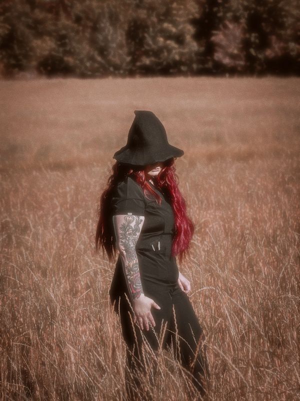 Woman with red hair and tattoos wearing a black witch hat in a field.