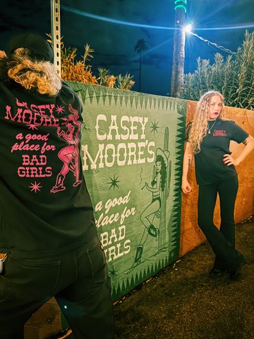 Two women modeling Casey Moore's themed shirts near a painted wall at night.