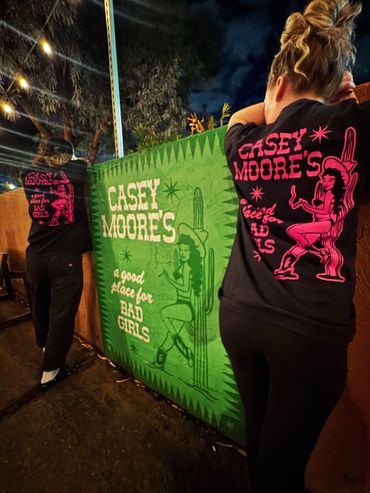Two people lean on a wall with 'Casey Moore's' shirts under a night sky.