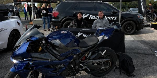 Two men sit behind a table at a motorcycle event with a sleek blue sports bike in front.