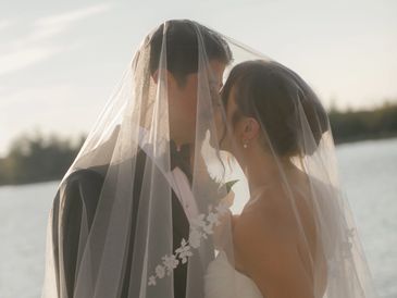 Bride and groom sharing a tender moment under a veil by the water.