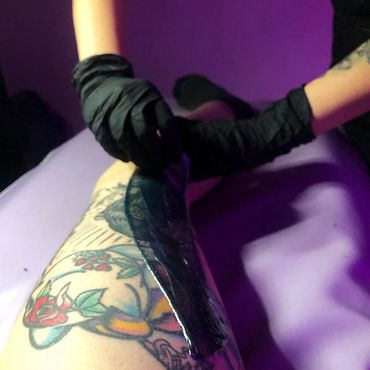 Person applying a black peel-off mask on tattooed leg wearing black gloves.