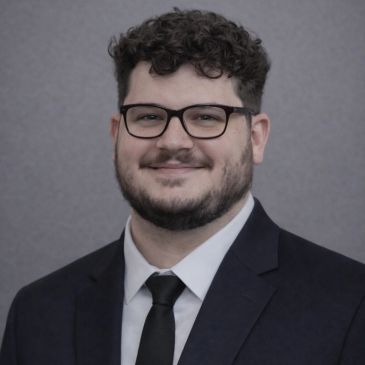 Smiling man with glasses in a suit and tie against a gray background.