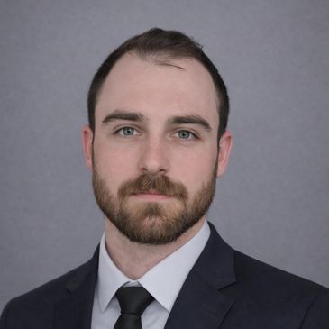 Young man in a dark suit and tie with a neutral expression.