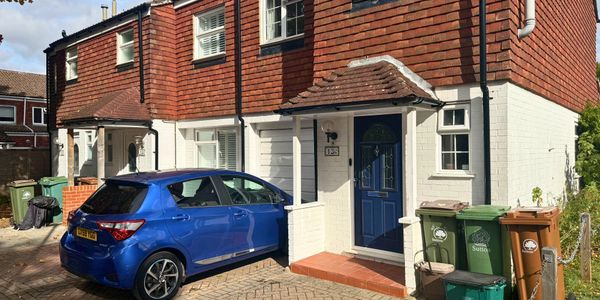 Blue car parked outside a red-brick and white house with bins.