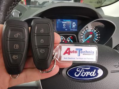 2 Ford remote proximity car keys in front of a steering wheel with instruments in background