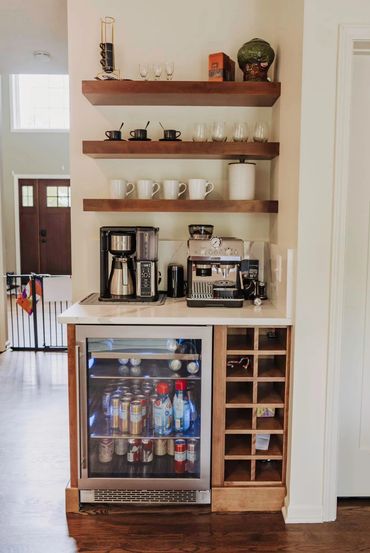 A stylish coffee station with machines, mugs, glasses, and a small fridge.