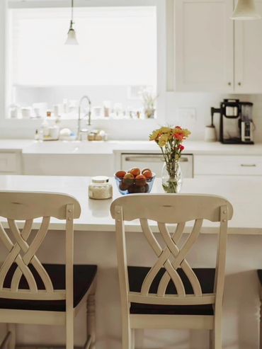 Bright kitchen with white chairs and a flower vase on the counter.