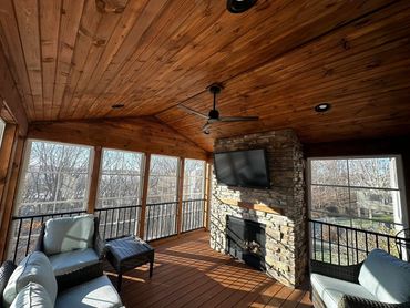 Cozy screened porch with stone fireplace and ceiling fan, surrounded by large windows.