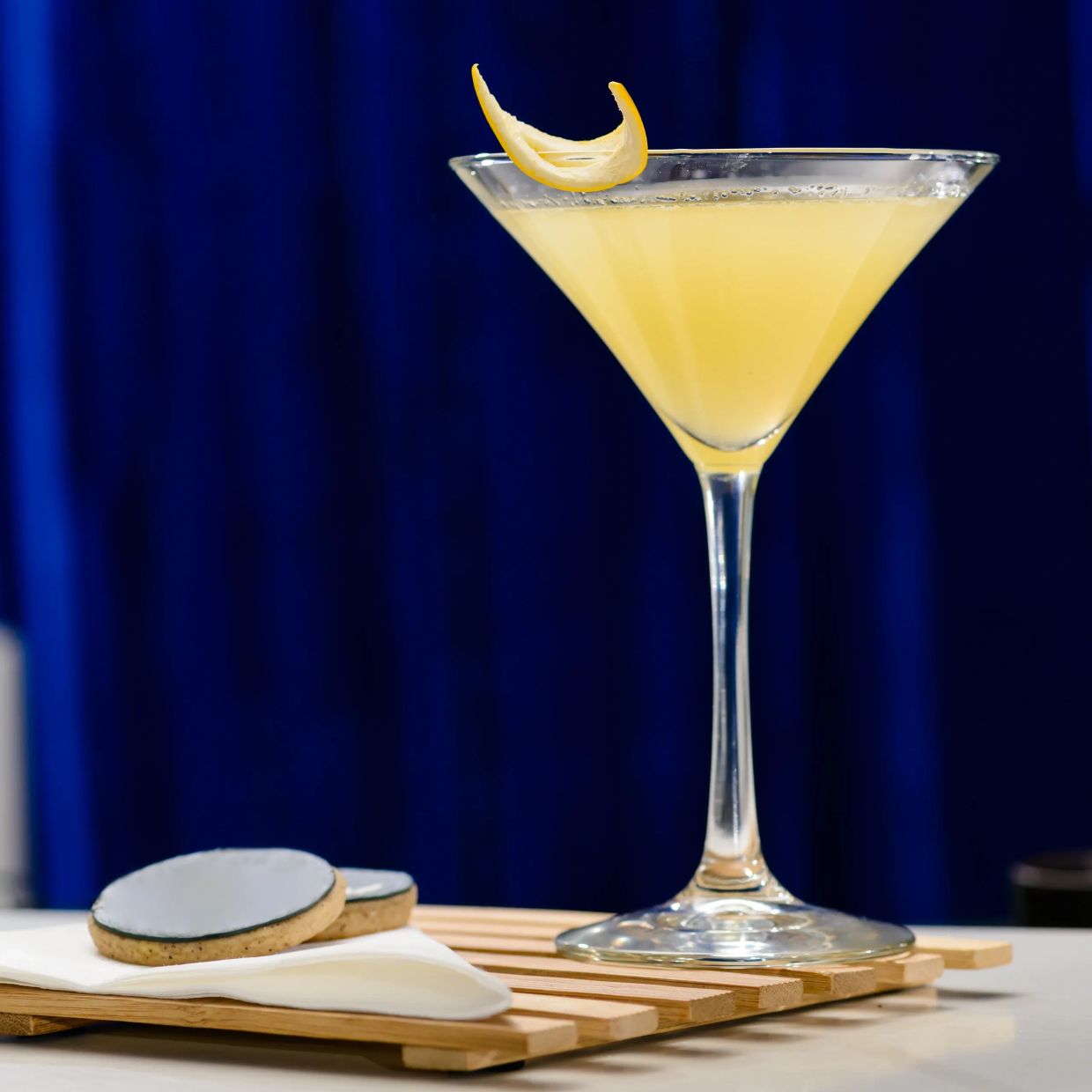 A yellow cocktail with a lemon twist and cookies on a wooden tray.