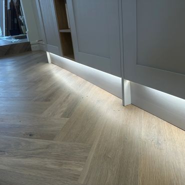 Under-cabinet lighting illuminates a modern kitchen floor with herringbone wood pattern.