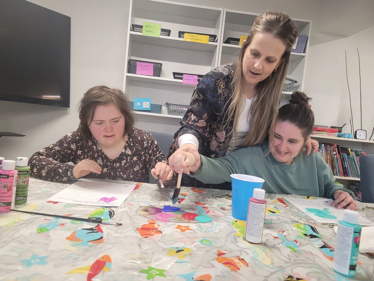 A woman helps two girls paint at a colorful table covered with art supplies.