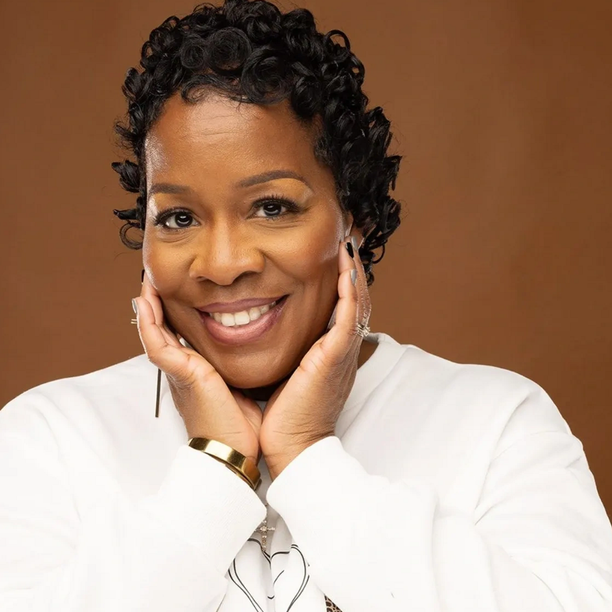 Smiling woman with curly hair posing with hands on her cheeks, wearing a white top.