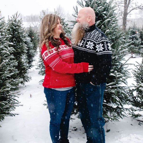 Couple in festive sweaters sharing a snowy moment among Christmas trees.