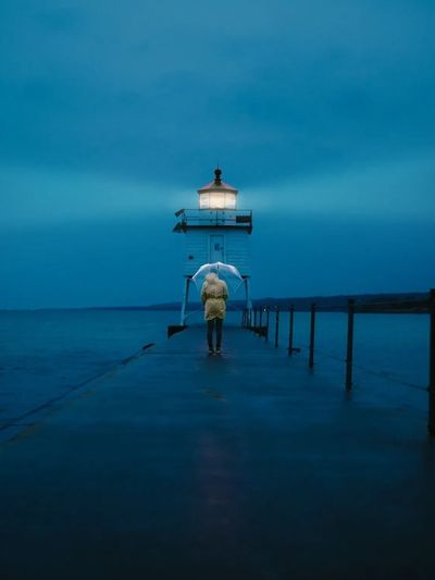 Person with umbrella walking toward a lit lighthouse at dusk over calm waters.