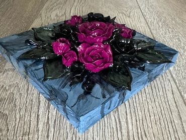 A dark, square artwork featuring deep purple and black roses surrounded by green leaves