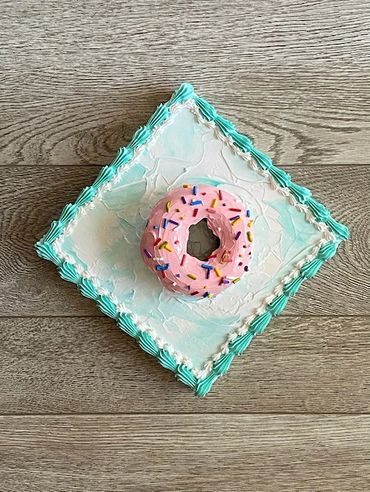 A chocolate donut with pink frosting and sprinkles, placed on a blue-iced square base.