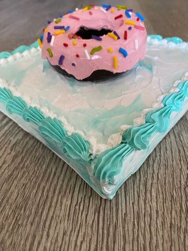 A close up of a chocolate donut with pink frosting and sprinkles, placed on a blue-iced square base