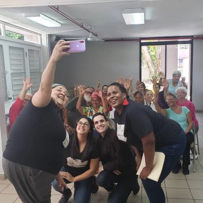A group selfie with smiling people and elderly participants waving in a community room.