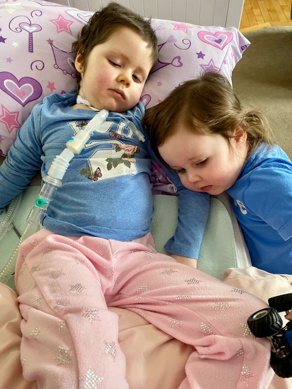 Two young children, one lying in bed with a medical tube, the other gently resting beside them.