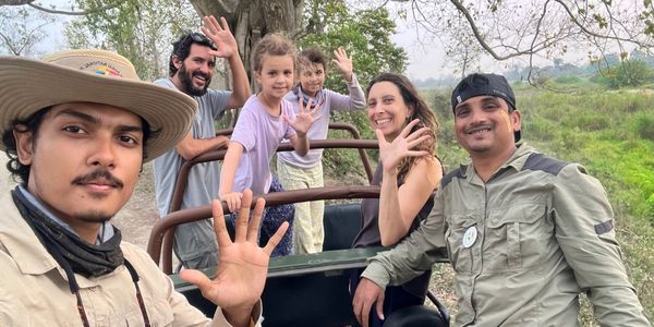 Group on a safari jeep waving at the camera in a green outdoor setting.