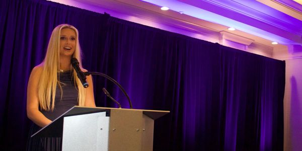 A woman with long blonde hair speaking at a podium with purple stage lighting.