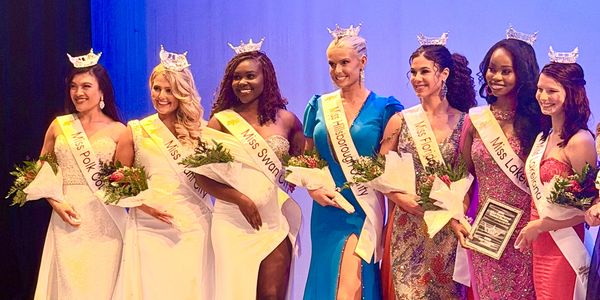 Seven women wearing crowns and sashes, holding flowers and awards on stage.
