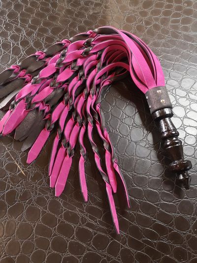 Pink and black leather flogger with a wooden handle on textured surface.