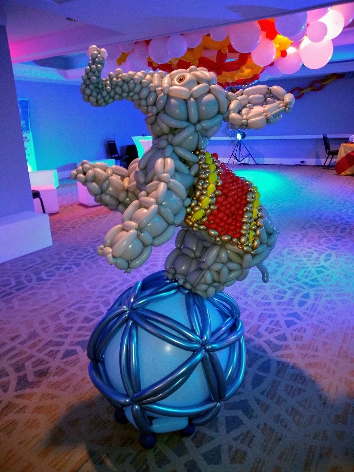A detailed balloon sculpture of an elephant balancing on a decorated blue ball indoors.