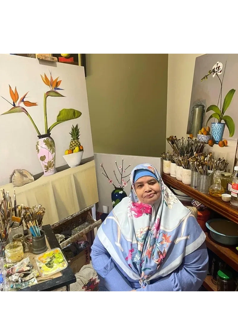 Woman in a floral headscarf sitting in an art studio surrounded by paintings and brushes.
