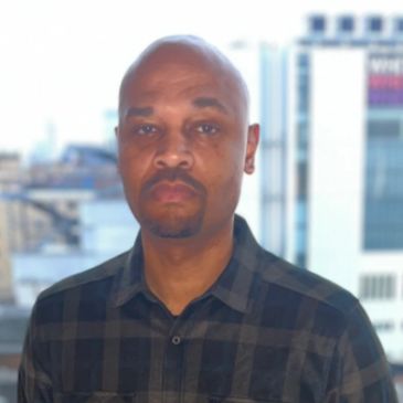 Man with shaved head and beard in a checkered shirt.