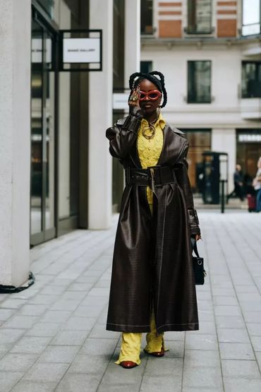 Fashionable woman in oversized brown leather coat and yellow outfit with red sunglasses.