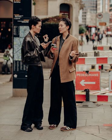 Two women discussing while holding smartphones on a city sidewalk.