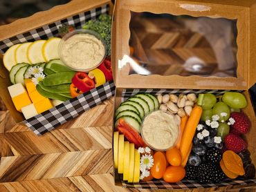 Two colorful snack boxes with fresh vegetables, fruits, cheese, nuts, and dip.