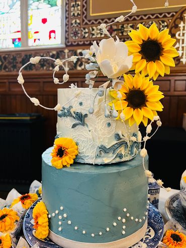 Sunflowers edible, white and blue wedding cake, two their. Set on blue and white dish at church.