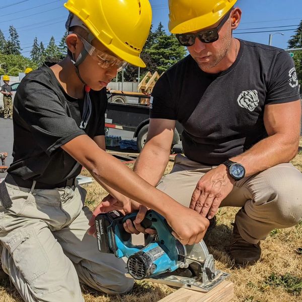 Adult teaching a young boy to use a saw, both wearing yellow helmets.