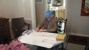 A man in a blue hat and suit looking at a large document on a table.