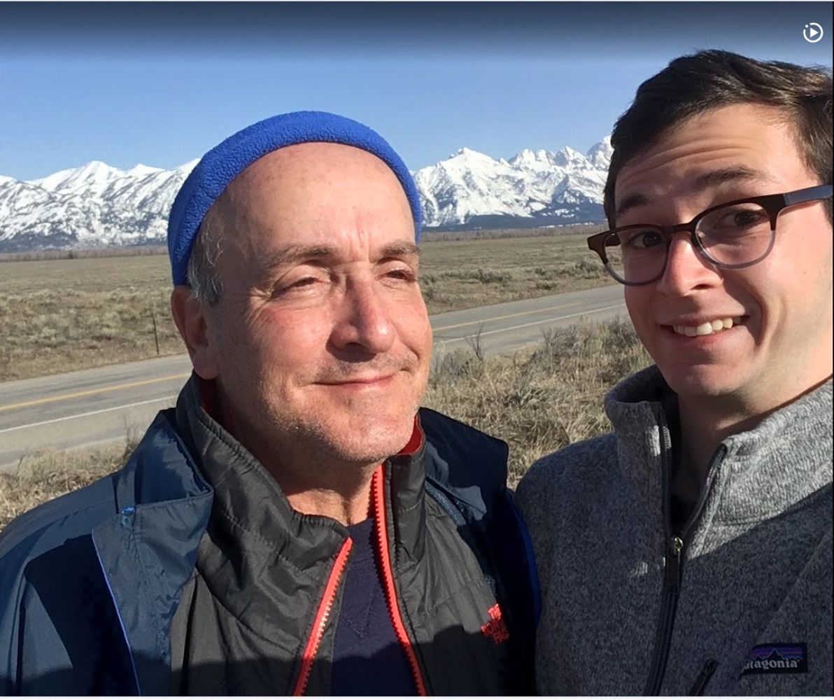 Two men smiling outdoors with snowy mountains in the background.