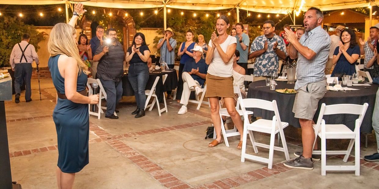 A woman speaks to an applauding audience at an outdoor event under string lights.