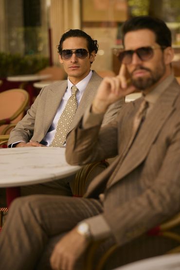 Two stylish men in suits and sunglasses sit at a cafe table.