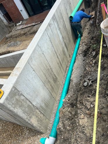 Worker installing a green drainage pipe beside a concrete wall.