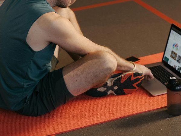 Man sitting on an orange yoga mat using a laptop while wearing athletic gear.