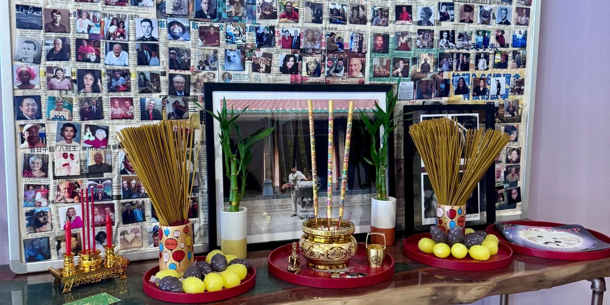 Altar with incense sticks, candles, and offerings in front of a photo collage and framed picture.