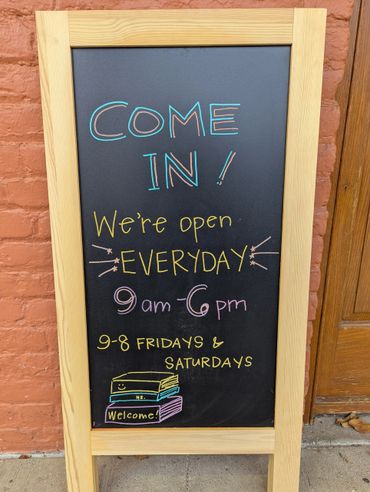 Chalkboard sign with colorful lettering showing store hours and welcome message.