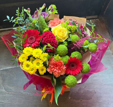A vibrant bouquet featuring a mix of red, yellow, orange, and green flowers wrapped in pink plastic.