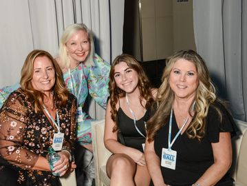 Four women smiling and posing indoors at a social event.