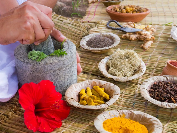 Hands grinding herbs in a stone mortar surrounded by various spices and a red hibiscus flower.