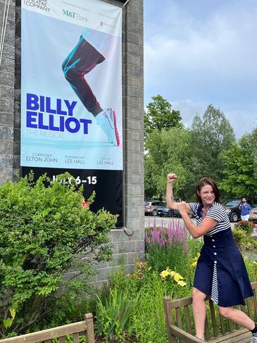 Woman posing energetically in front of a Billy Elliot poster.
