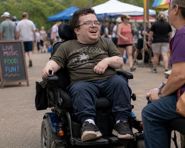 Man in wheelchair smiling and talking to a seated person at an outdoor event.