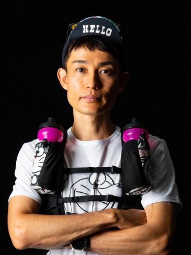 Man wearing a cap and hydration vest with water bottles, arms crossed against black background.